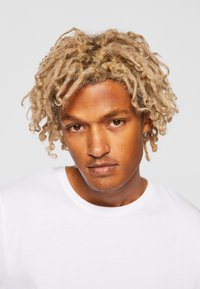 Porträt einer Person mit kurzen, lockigen blonden Haaren, die ein schlichtes weißes T-Shirt mit rundem Ausschnitt und glatter Stoffoberfläche trägt.