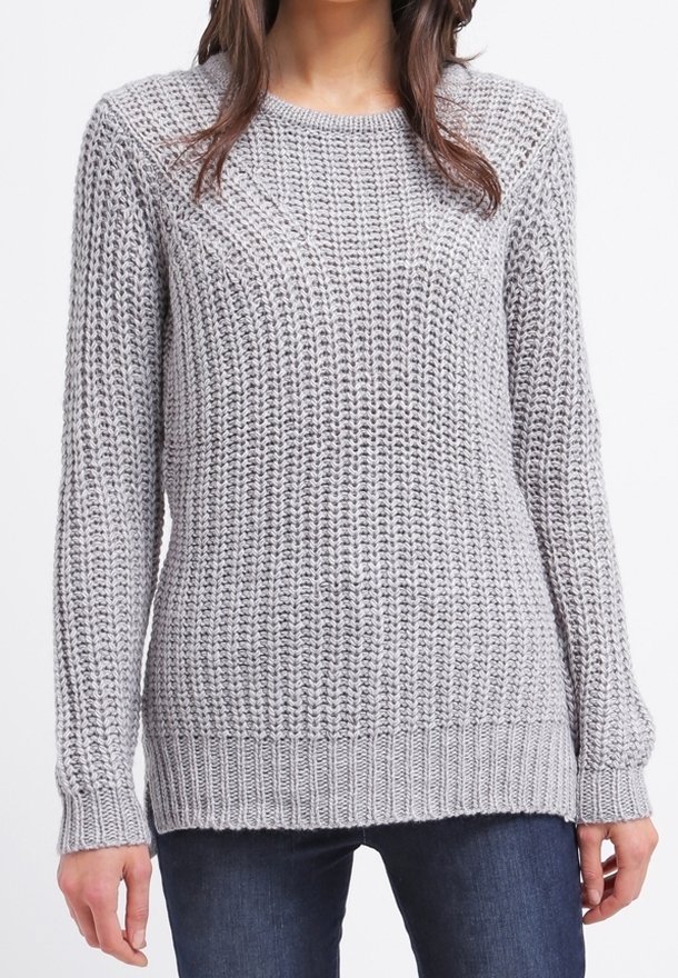 Femme portant un pull gris clair en grosse maille avec des manches longues et des poignets côtelés, associé à un jean bleu foncé.