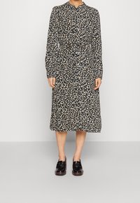 Donna che indossa un vestito a maniche lunghe, lungo fino al ginocchio, con stampa astratta nera, beige e bianca, fermato in vita, e mocassini neri lucidi.