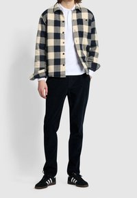 Chemise en flanelle à carreaux crème et bleu marine, avec boutons et deux poches poitrine, associée à un pantalon foncé et des baskets noires à rayures blanches.