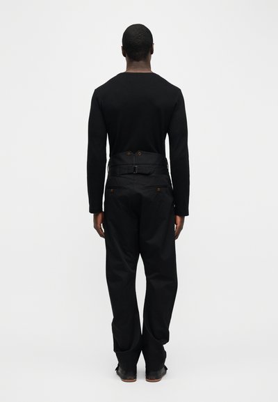 Άνδρας που φοράει μαύρο πουκάμισο μακρυμάνικο και ψηλόμεσο μαύρο παντελόνι με τσέπες στο πίσω μέρος με κουμπιά, στέκεται γυρισμένος με την πλάτη σε λευκό φόντο.