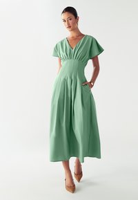 Vestido midi verde con mangas cortas y acampanadas, cintura fruncida y bolsillos laterales. Tejido suave con una silueta fluida, combinado con tacones estampados.