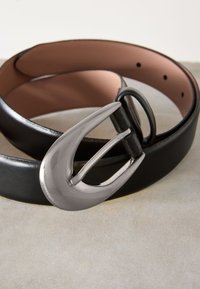 Črni usnjeni pas s srebrno, polirano, preveliko sponko. Notranja stran je gladek rožnat material z več luknjami za prilagajanje.