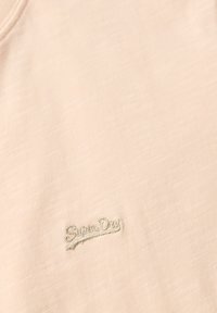 T-shirt rose clair avec une texture douce, présentant un petit logo "Superdry" brodé dans une couleur contrastante. Design simple et minimaliste.