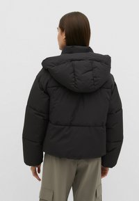 Sort puffervest med hætte, der har en kasseformet silhouette, quilted tekstur og synlige sømme på ryggen. Bæres med løse beige cargo-bukser.