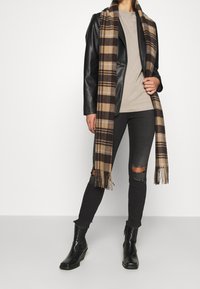 Svart skinnjacka, beige T-shirt, rutig scarf i brunt och tan, slitna svarta jeans och svarta ankelskor med en kraftig klack.