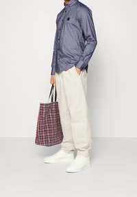 Camisa azul marino con botones, pantalones beige, zapatillas blancas, sosteniendo una bolsa tote a cuadros roja y negra con asas negras. Atuendo sencillo y casual.