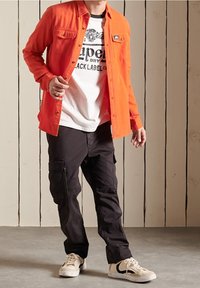 Oranssi nappipaita taskuilla valkoisen graafisen t-paidan päällä. Mustat cargo-housut vetoketjuilla ja taskuilla. Valkoiset tennarit.