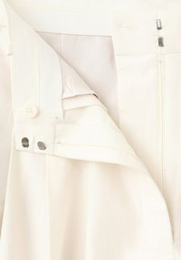 Gros plan sur un pantalon de couleur crème avec une fermeture éclair, un bouton et des agrafes doubles partiellement ouvertes.