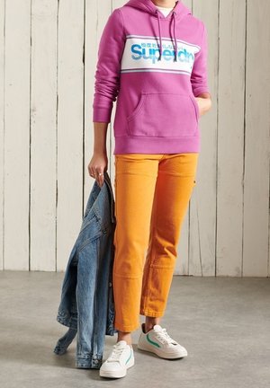 Sudadera con capucha rosa con un logo blanco y azul, pantalones ajustados naranjas, chaqueta vaquera en la mano y zapatillas blancas con detalles verdes.
