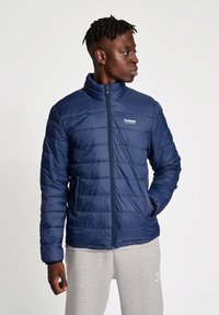 Marinblå isolerad puffjacka med horisontell sömnad, ståkrage och dragkedjestängning. Logotyp på övre vänstra sidan.