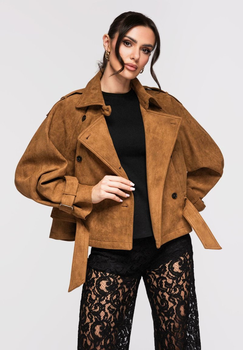 Femme aux cheveux foncés portant une veste en daim marron sur un haut noir et un pantalon en dentelle noire, posant devant un fond clair uni.
