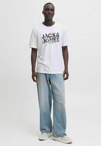 Balta īsām piedurknēm T-kreklu ar krāsainu "JACK & JONES" grafiku uz priekšpuses, saskaņots ar gaiši ziliem platām kājām džinsiem un baltām sporta apaviem.