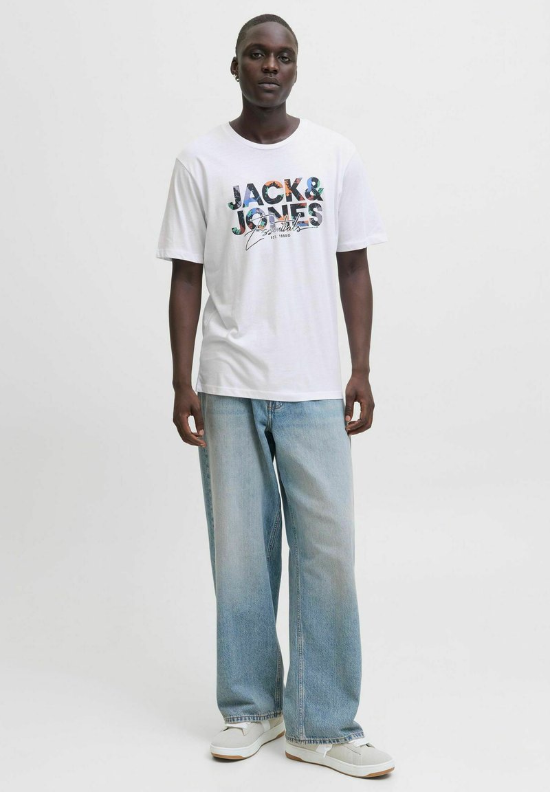 Balta īsām piedurknēm T-kreklu ar krāsainu "JACK & JONES" grafiku uz priekšpuses, saskaņots ar gaiši ziliem platām kājām džinsiem un baltām sporta apaviem.