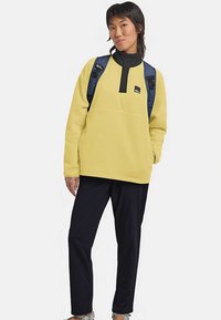 Yellow fleece pullover with a black collar and button placket, paired with dark pants and a blue backpack. Gray shoes are also visible.