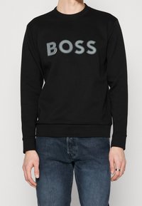 Sudadera negra con un prominente logo "BOSS" en gris en el pecho, escote redondo, puños y dobladillo acanalados. Combinada con jeans azules.