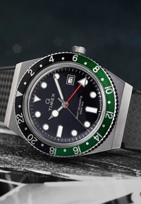 Montre Timex Q avec un boîtier en acier inoxydable, un cadran noir, une lunette verte, des index horaires lumineux et un bracelet en caoutchouc texturé.