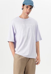 Fialové oversized tričko s textem "LOVERS IN" v bílé barvě. Krátké rukávy a kulatý výstřih. V kombinaci se světle khaki kalhotami.
