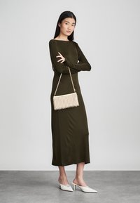 Olívazöld, hosszú ujjú ruha egyenes sziluettel. Gyöngyökkel díszített borítéktáskával arany láncon és fehér magassarkú cipővel kiegészítve.