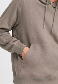 Close-up of a person wearing a gray hoodie with a front pocket, showing a hand resting in the pocket and part of the sleeve.