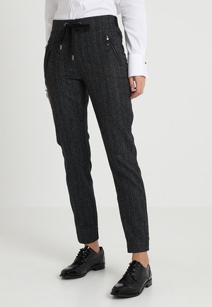 Kobieta ubrana w ciemnoszare spodnie w cienkie paski z frontowymi kieszeniami na zamek błyskawiczny, białą koszulę oraz czarne eleganckie buty sznurowane.