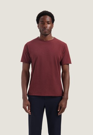 Man met kort krullend haar die een effen donkerrode korte mouwen T-shirt en een donkere broek draagt, staand tegen een lichte achtergrond.
