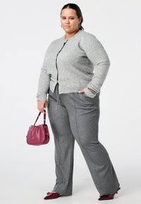 Vrouw die een grijze cardigan met pareldetails draagt, grijze pantalon met krijtstrepen, een bordeauxrode handtas vasthoudt en bijpassende bordeauxrode puntige schoenen draagt.