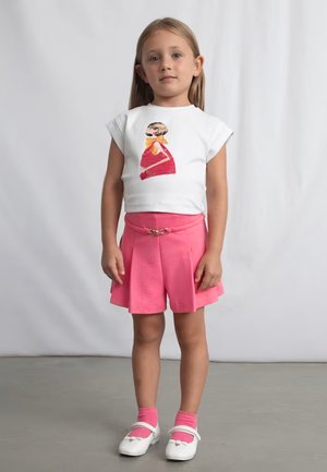 Ragazza giovane in piedi all'interno, indossa una maglietta bianca con una grafica, pantaloncini rosa con cintura, calzini rosa e scarpe bianche.