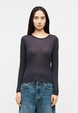 Νεαρή γυναίκα με μακριά σκούρα μαλλιά φοράει διάφανο σκούρο μακρυμάνικο τοπ και ψηλόμεσο μπλε τζιν, στέκεται μπροστά σε ένα απλό φόντο.