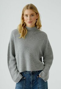Lichtgrijze, cropped pullover trui met een hoge hals en lange mouwen, gemaakt van een zacht breimateriaal. Gecombineerd met blauwe denim onderkleding.