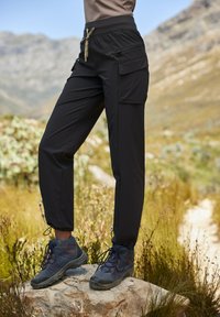 Schwarze Cargo-Hosen mit elastischem Bund, seitlichen Taschen und verstellbaren Knöchelbündchen, kombiniert mit dunklen Wanderschuhen auf steinigem Terrain.