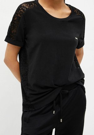 Kobieta w czarnym krótkim rękawie z koronkowymi panelami na ramionach i bokach, w połączeniu z czarnymi spodniami ze ściągaczem.