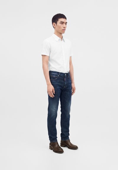 Homme debout portant une chemise blanche à manches courtes boutonnée, un jean bleu foncé et des bottes en cuir marron sur un fond clair uni.