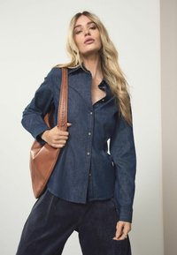 Tmavé deninové košele s dlhými rukávmi, v kombinácii s námorníckymi kordovými nohavicami. Outfit dopĺňa hnedá kožená taška s pletenou rukoväťou.