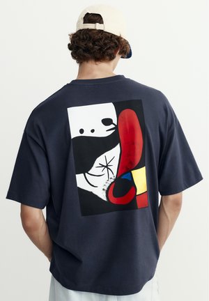 Osoba nosząca granatowy T-shirt z abstrakcyjnym czerwonym, czarnym, białym, żółtym i niebieskim wzorem na plecach oraz beżową czapkę z daszkiem.