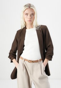 Smeđi sakou prilagođenog kroja s glatkom teksturom, nosi se preko bijele košulje i uparuje s bež hlačama visokog struka i smeškim remenom.