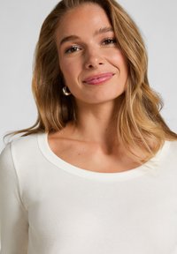 Jonge vrouw met lange blonde haren, draagt zilveren creolen en een witte geribbelde top, glimlacht zachtjes tegen een effen achtergrond.