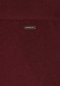 Bordeaux gebreide stof met ribbeltextuur; heeft een zilver logo-tag die "DREMASTER" op het oppervlak leest.