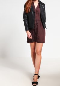 Kobieta ubrana w czarną skórzaną kurtkę na brązową sukienkę do kolan z wiązaniem w talii, w połączeniu z czarnymi sandałami na wysokim obcasie z kostkowym zapięciem.