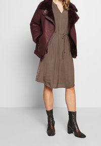 Maroons shearling-lined leather jacket over a brown dotted dress with a tie waist, paired with high-heeled, patterned ankle boots.
