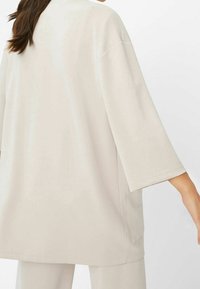 Světle béžový oversized top vyrobený z hladké látky. Má široké rukávy, zaoblený výstřih a boční rozparek na lemu pro snadnější pohyb.