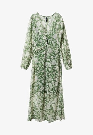 Vrouw draagt een groene bloemenprint midi-jurk met lange mouwen en een vooraan openstaande split, gecombineerd met zwarte enkellaarsjes met metalen neuzen.