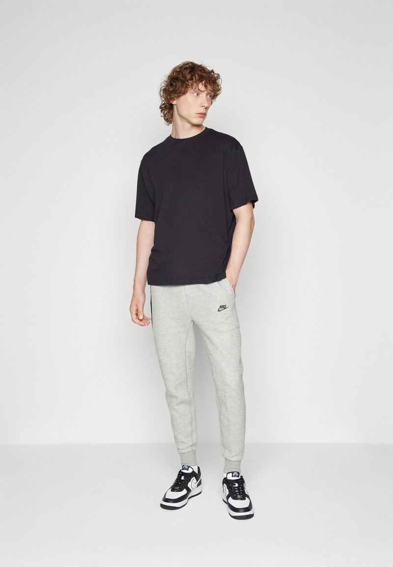 Schwarzes T-Shirt mit kurzen Ärmeln, kombiniert mit hellgrauen Sweatpants mit Nike-Logo. Abgerundet wird das Outfit mit schwarz-weißen Sportschuhen.