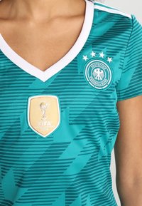 Turkoosi jalkapallopaita, jossa on valkoinen V-pääntie, vaakasuorat raidat ja embleemit: FIFA World Cup 2014 -merkki ja Deutscher Fussball-Bund -logo.