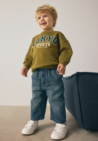 Oliwkowy sweter z napisem "TOKYO SPORTS", w zestawie z niebieskimi dżinsami i białymi trampkami na rzepy. Krótkie kręcone włosy i radosny wyraz twarzy.