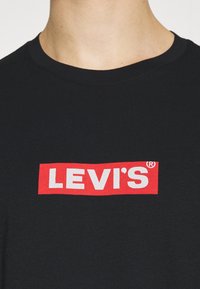 Zbliżenie na osobę w czarnej koszulce z czerwonym prostokątnym logo Levi's w białych literach na klatce piersiowej.