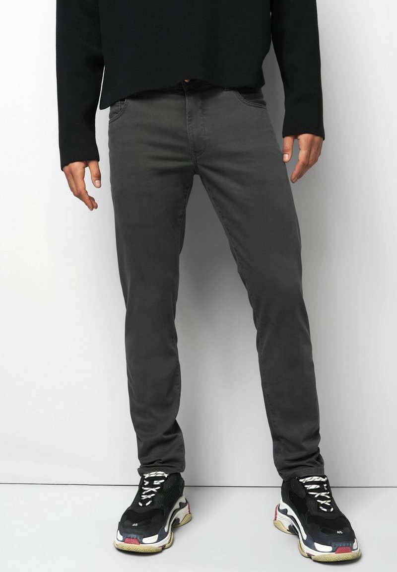Man in een donkergrijze broek, een zwart langetermijnshirt en zwarte chunky sneakers met witte en rode accenten die tegen een witte achtergrond staan.