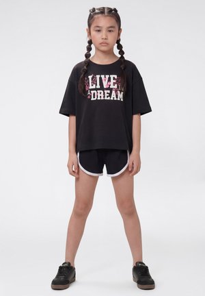 Ragazza giovane con capelli intrecciati, in piedi di fronte, indossa una t-shirt nera "Live a Dream", pantaloncini neri con bordo bianco e sneakers nere.