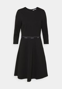 Černé šaty s kulatým výstřihem a tříčtvrtečními rukávy; mají vypasovaný pas s kontrastním páskem s logem Calvin Klein. Hladká tkanina.