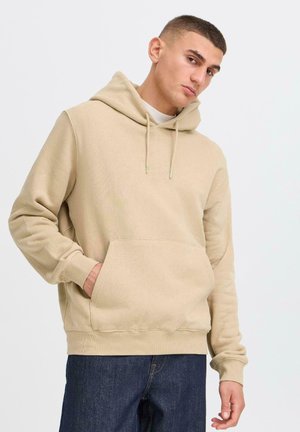 Young man wearing a beige hoodie with a front pocket, standing casually with his left hand in the pocket, against a white background.